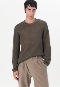 Pulover tricotat maro cu un model texturat și manșete cu zimți, asociat cu pantaloni plisati bej deschis. Croi casual, design cu gât rotund.