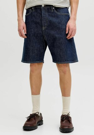 Shorts en jean bleu foncé pour hommes portés avec des chaussettes blanches et des chaussures en cuir marron à lacets, visibles de la taille jusqu'aux pieds.