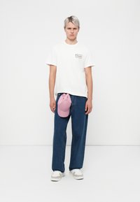 White T-shirt with a logo patch, loose blue denim pants, pink cap resting on the waistband, and white sneakers with gray accents.