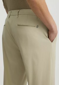 Pantalones beiges con una textura suave, que presentan bolsillos traseros con aberturas horizontales y un detalle de logo cerca de la cinturilla.
