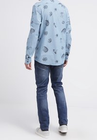 Man in blauwe jeans, witte sneakers en een lichtblauw shirt met lange mouwen en donkere botanische prints, staand met de rug naar de camera op een witte achtergrond.