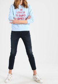 Osoba v světle modrém svršku s červeným textem "ŽÍT JE TANČIT," černých džínách a bílých teniskách stojící se zkříženýma rukama.