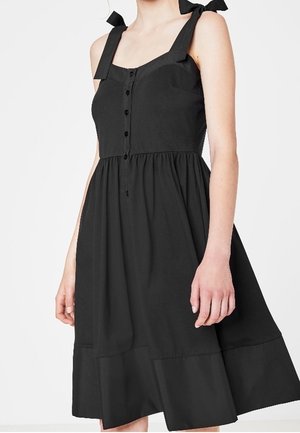 Schwarzes Baumwollkleid mit breiten Trägern, Knopfleiste vorne und geraffter Taille. Verfügt über einen ausgestellten Rock und einen mehrstufigen Saum. Bindebänder an den Trägern.