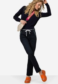 Pantalones de mezclilla oscura con cordón blanco, combinados con unas sandalias de plataforma naranjas. El modelo lleva una blusa azul marino y sostiene un bolso cruzado beige.