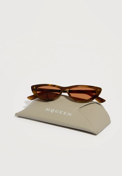 Καφέ γυαλιά ηλίου με σχήμα γάτας που ακουμπούν σε μια μπεζ θήκη γυαλιών με την επωνυμία McQueen, σε λευκό φόντο.