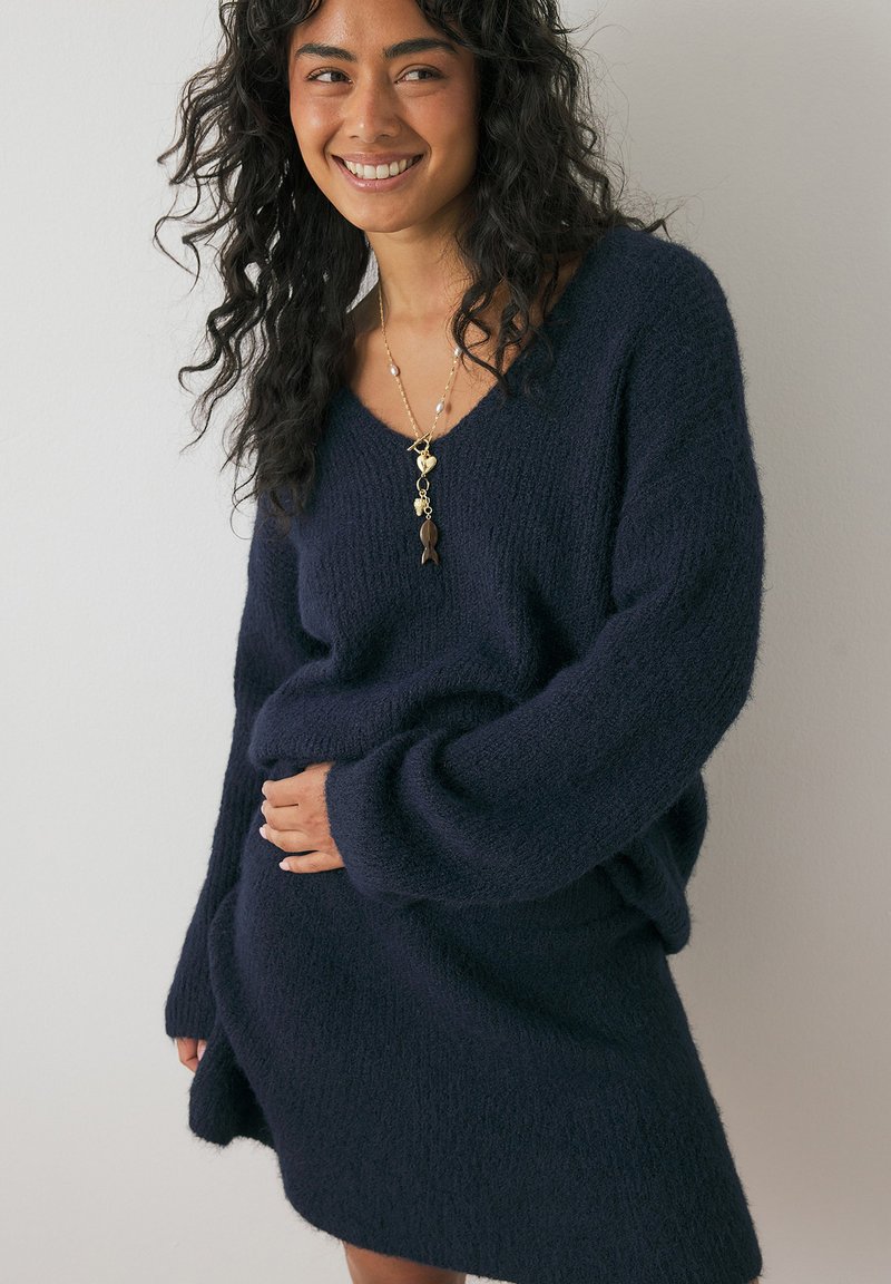 Mörkblå stickad klänning i en texturerad, luftig väv, med v-ringning och långa ärmar; bärs avslappnat med ett lager av halsband.