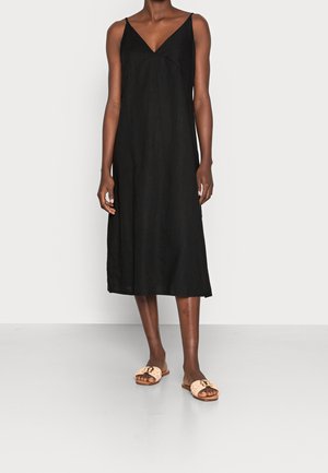 Femme portant une robe midi noire sans manches avec de fines bretelles et des sandales beige à enfiler avec des boucles décoratives.
