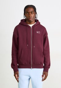 Bordeaux zip-up hoodie med en snøre hætte og frontlommer. Har et hvidt Nike-logo på venstre bryst. Glat tekstur.
