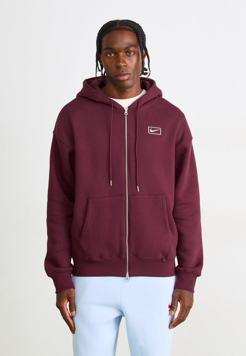 Burgunderfarget zip-opp hettegenser med snorhette og frontlommer. Har en hvit Nike-logo på venstre bryst. Glatt tekstur.