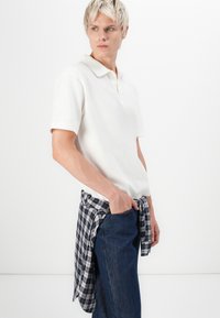 White polo shirt, short sleeves, paired with dark blue denim jeans. A checkered shirt tied around the waist features navy and white patterns.