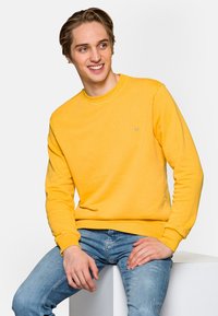 Żółta bluza z długim rękawem i okrągłym dekoltem. Miękka tkanina, ściągacze przy rękawach oraz subtelny detal logo na piersi. Noszona z niebieskimi dżinsami.