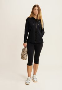 Felpa nera con zip e dettagli dorati, abbinata a leggings capri neri. Tiene una borsa beige con un motivo testurizzato multicolore. Sneakers argentate.