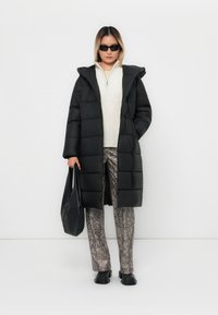 Sort pufferjakke med hætte, lange ærmer og firkantet syning. Bæres over en hvid sweater og mønstrede beige slangeprint bukser. Sort taske.