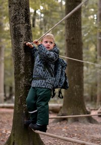 Barn i grå jakke med mønster, grønne bukser og sorte støvler klatrer i et træ og holder fast i et tov i en skov.