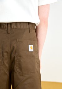 Pantaloni din bumbac maron cu două buzunare mari la spate și un patch proeminent cu logo-ul Carhartt. Croi ajustat, cusături vizibile și o textură durabilă.