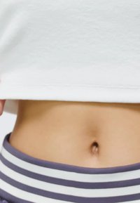 Vit crop top hålls ovanför ett högmidjebelts med grå och vita ränder. Slät, mjuk tygtextur synlig, som visar upp magen.