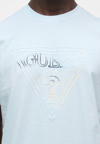 Άνδρας που φοράει ανοιχτό μπλε μπλουζάκι με ανάγλυφο λογότυπο Guess U.S.A. που απεικονίζει ένα τρίγωνο και ένα ερωτηματικό στο στήθος.