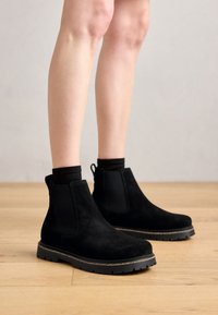 Cizme negre din suede până la glezne, cu panouri laterale elastice, talpă din cauciuc texturat și fâșie de prindere. Culoare uniformă cu detalii minime.