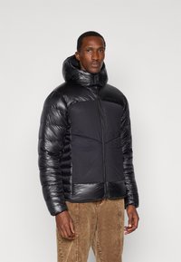 Homme portant une doudoune noire matelassée avec un col haut et une capuche, associé à un pantalon en velours côtes chocolat, se tenant contre un fond uni.