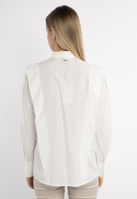 Blusa bianca a maniche lunghe con colletto alto, dettagli plissettati sulle spalle e polsini con bottoni, caratterizzata da una texture liscia e un sottile accento con logo.