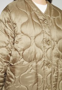 Veste matelassée beige avec un quilting abstrait aux coutures courbes, boutons à l'avant et poches latérales, portée sur un haut blanc.