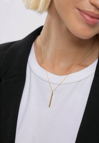 Auksinis pakabukas ant grandinėlės su elegantišku vertikaliu juostos dizainu. Dėvimas ant baltos palaidinės, derinamas su juodu švarku. Minimalistinis ir elegantiškas.