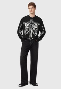 Zwarte gebreide trui met een witte skeletprint op de voorkant en mouwen, gecombineerd met zwarte wijde jeans en zwarte schoenen.