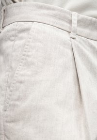 Pantalon beige clair en tissu texturé, avec un pli sur le devant, une ceinture plate et des poches latérales.