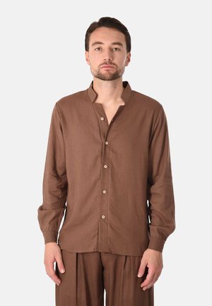 Uomo con capelli corti e scuri e barba, che indossa una camicia marrone a maniche lunghe con collo alla coreana e pantaloni marroni abbinati, in piedi immobile.
