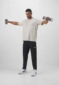 Lys beige t-skjorte og svarte joggebukser med hvit Adidas-logo; holder sølv manualer; står med armene utstrakt, idrettssko.