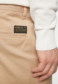 Pantalons en coton beige avec une finition texturée. Présente une étiquette sombre portant la mention "REPLAY" près de la poche droite. Un pull en coton blanc à côtes est visible.