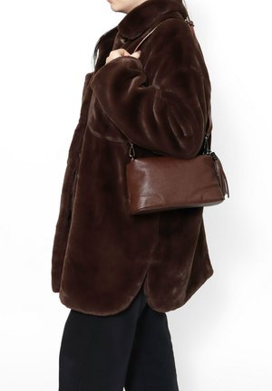 Persona che indossa un cappotto in pelliccia ecologica marrone scuro, porta una piccola borsa a tracolla in pelle marrone, mostrata di profilo su sfondo bianco.