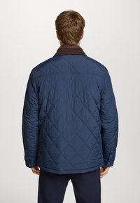 Chaqueta acolchada de color navy con un patrón de diamantes, que cuenta con un cuello de canalé marrón y botones decorativos en los puños. Textura suave, diseño práctico.