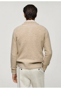 Maglione beige a maglia con colletto, orlo a costine e dettagli di texture. Indossato in modo casual su pantaloni chiari.