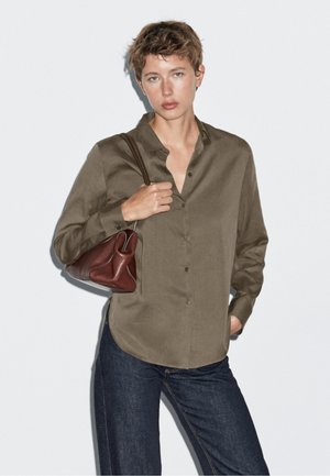 Camicia verde oliva con colletto e chiusura a bottone, abbinata a jeans di denim scuri e una piccola borsa di pelle marrone, tenuta nella mano destra.