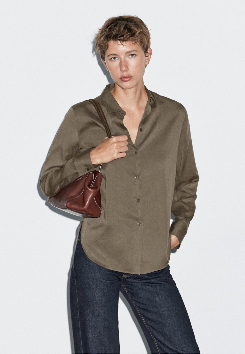 Camicia verde oliva con colletto e chiusura a bottone, abbinata a jeans di denim scuri e una piccola borsa di pelle marrone, tenuta nella mano destra.
