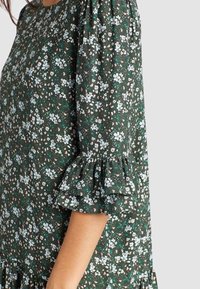 Vestido floral con fondo verde, que presenta flores blancas y rosas, mangas abullonadas y volantes en los bordes del dobladillo y las mangas.