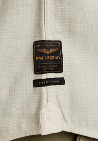 Tissu beige avec deux étiquettes noires, l'une affichant un aigle doré et le texte "PME Legend American Classic Tested at High Altitude", et l'autre avec "Type N° C-46".