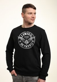 Sudadera negra con un gráfico circular que presenta alas y el texto "LYNYRD FREEBIRD 1973". Puños y dobladillo acanalados, textura suave.