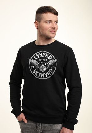 Sort sweatshirt med et cirkulært grafisk motiv med vinger og teksten "LYNYRD FREEBIRD 1973." Ribbede manchetter og søm, blød tekstur.