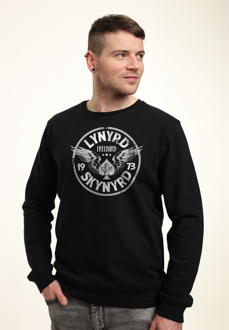 Sudadera negra con un gráfico circular que presenta alas y el texto "LYNYRD FREEBIRD 1973". Puños y dobladillo acanalados, textura suave.