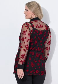Blusa nera trasparente con ricami floreali rossi, maniche lunghe e polsini con bottone; presenta un bordo in pizzo lungo il colletto.