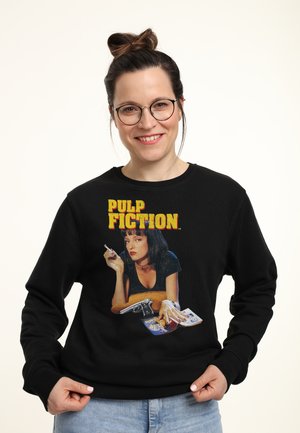 Kvinde med briller og håroppsutning iført sort sweatshirt med Pulp Fiction-grafik, der viser en kvinde med en cigaret, pistol og penge.