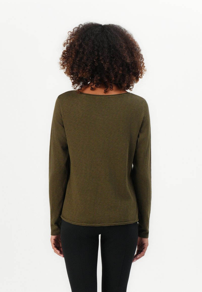 Kobieta z kręconymi włosami, ubrana w oliwkowo-zieloną koszulę z długim rękawem i czarne spodnie, stoi przodem do białego tła, z rękami opuszczonymi wzdłuż ciała.