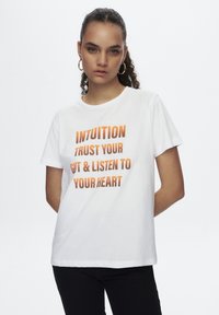 T-shirt en coton blanc avec une typographie orange audacieuse indiquant "INTUITION FAIS CONFIANCE À TON INSTINCT ET ÉCOUTE TON COEUR." Coupe classique, manches courtes.