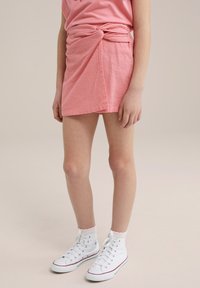 Rozā teksturēta mini svārku ar savītu mezglu dizainu jostasvietā, pārī ar baltiem augstiem sporta apaviem un potīšu zeķēm.