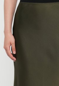 Olive green satin skirt with a smooth texture, featuring a black elastic waistband. Hand placed on the fabric, highlighting its flow.