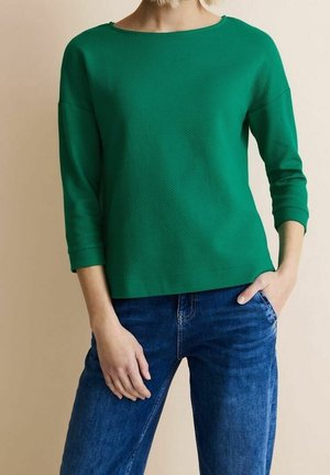 Kobieta ubrana w zielony, teksturowany top z długim rękawem i niebieskie dżinsy z jedną ręką w kieszeni na beżowym tle.