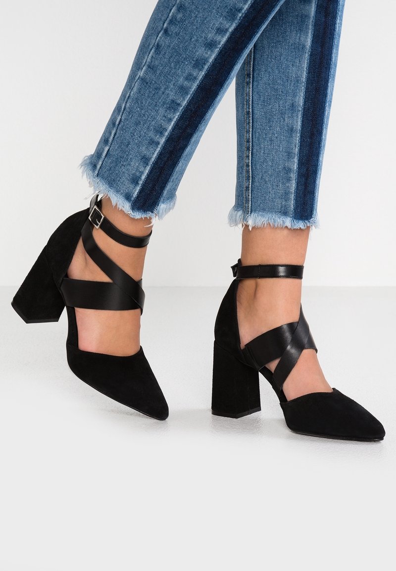 Pieds portant des talons blocs en daim noir avec des brides croisées à la cheville, associés à un jean bleu court avec une bande latérale foncée et un ourlet effiloché.