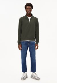 Dunkelgrüner Reißverschluss-Sweatshirt mit hohem Kragen, kombiniert mit hellblauen Jeans in gerader Form und grauen gemusterten Sneakers.